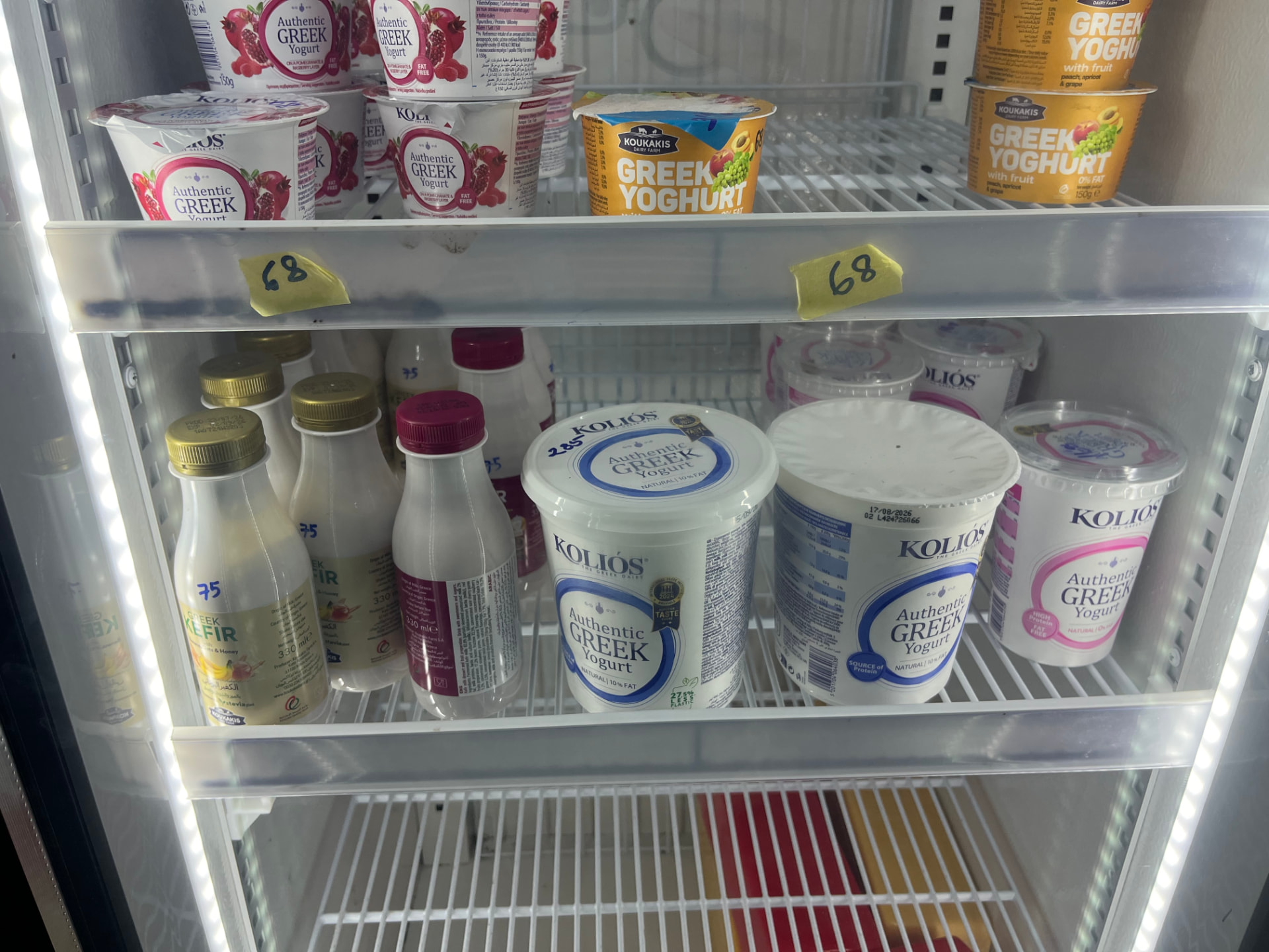 A refrigerated shelf of Greek yoghurt tubs with handwritten price tags in a Seychelles supermarket.