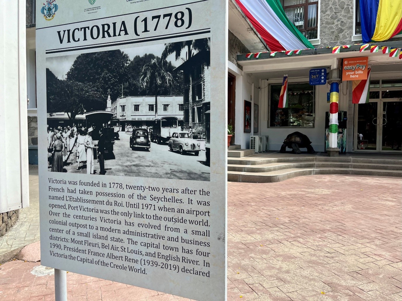 A commemorative plaque reading Victoria (1778) in the center of Victoria, Seychelles, next to a storefront.